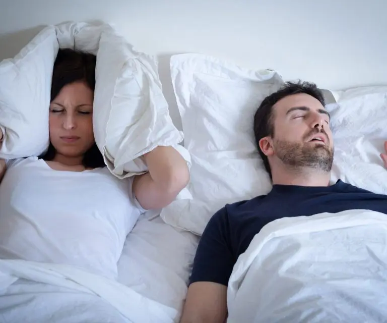 Mann und Frau im Bett; er schnarcht, sie hält sich das Kissen über die Ohren